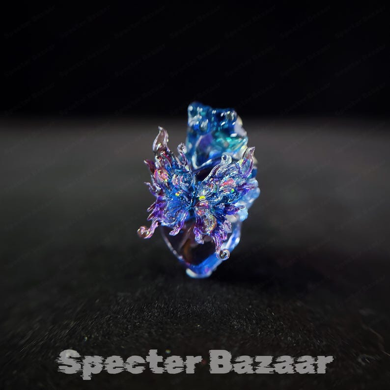 May include: A handcrafted, iridescent blue and purple glass ring with a floral design. The ring has a unique, artistic shape and is set against a dark background. The text "Specter Bazaar" is at the bottom.