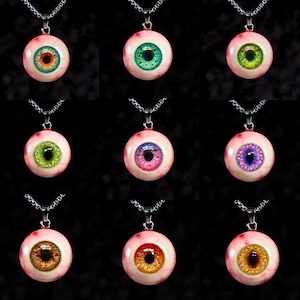 Puede incluir: Nueve collares con colgantes con diseños de globos oculares realistas. Cada colgante tiene un color de iris diferente, incluyendo verde, azul, marrón y morado, sobre un fondo blanco rosado. Los colgantes cuelgan de collares de cadena plateada.