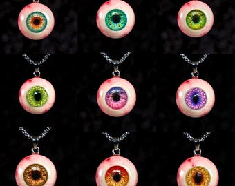 May include: Nine pendant necklaces featuring a realistic eyeball design. Each pendant has a different iris color, including green, blue, brown, and purple, set against a pinkish-white background. The pendants hang from silver-colored chain necklaces.