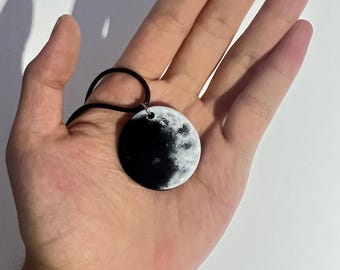 May include: A black and white moon pendant necklace with a black cord. The moon pendant is painted with a realistic depiction of the moon's surface.