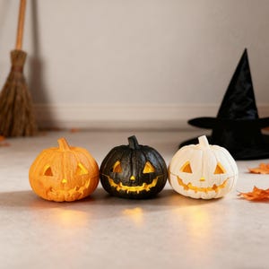 Puede incluir: Tres calabazas de Halloween iluminadas en naranja, negro y blanco. Cada calabaza tiene una cara tallada y una luz cálida en su interior. Un sombrero de bruja negro y una escoba sugieren un tema de Halloween.