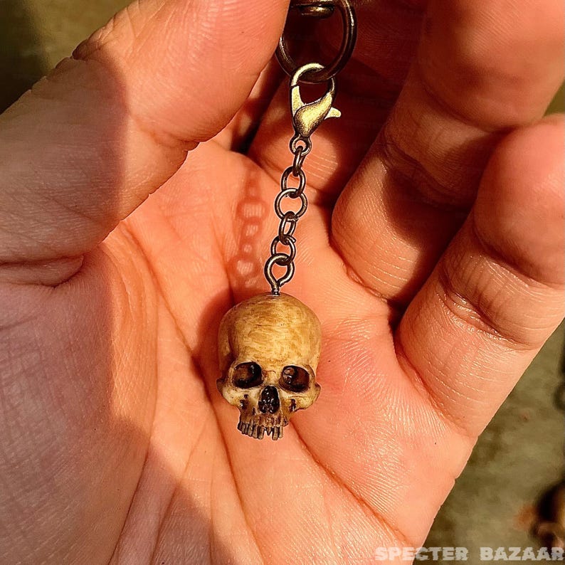 May include: A wooden skull keychain with a gold chain and clasp. The skull is detailed with eye sockets, a nose, and teeth.