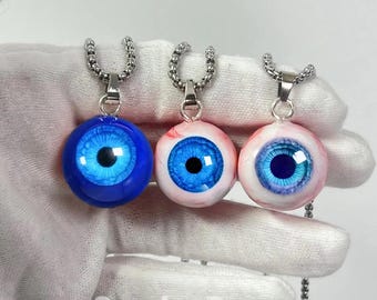 May include: Three eyeball pendants with blue irises and black pupils hang from silver chains. One pendant is solid blue, while the others are white and pink with red veins. The pendants are displayed against a white background.