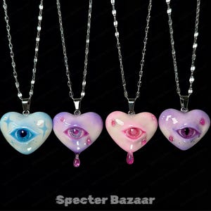 May include: Four heart-shaped pendants on silver-toned chains, each featuring a central realistic eye. The colors include light blue with star accents, purple with pink drips, soft pink with pink drips, and deep purple with pink drips.