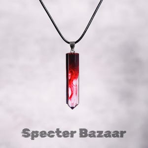 Puede incluir: Un collar con colgante con un colgante rectangular en forma de cristal. El colgante tiene un diseño de remolino rojo y blanco y cuelga de un cordón negro. El texto "Specter Bazaar" está en la parte inferior.