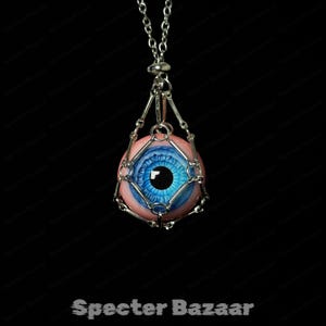 May include: A silver pendant necklace with a detailed eyeball design. The eye has a blue iris and is set in a pink and silver cage, hanging from a silver chain. The text "Specter Bazaar" is at the bottom.