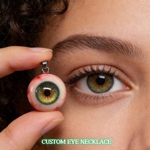 Puede incluir: Un collar con colgante con un diseño de ojo realista. El ojo tiene un iris verde y amarillo, venas rojas y una fianza plateada. El texto "CUSTOM EYE NECKLACE" se muestra en verde. El collar se sostiene cerca de un ojo.