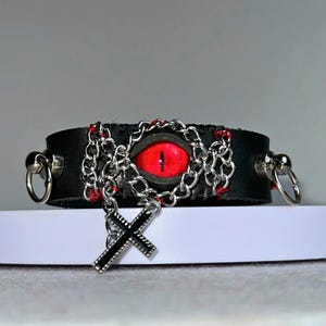 Puede incluir: Pulsera de cuero negro con un ojo rojo, una cadena plateada y un dije de cruz plateada.
