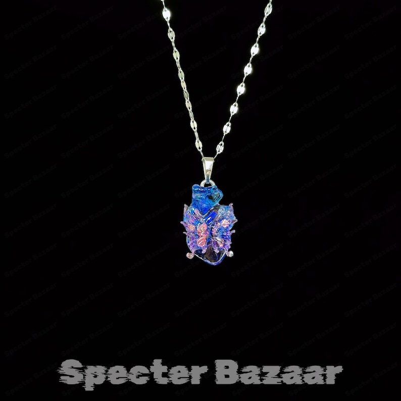 May include: A silver necklace with a pendant featuring a blue and purple design. The pendant has a butterfly-like shape with intricate details. The necklace is displayed against a black background. The text "Specter Bazaar" is at the bottom.