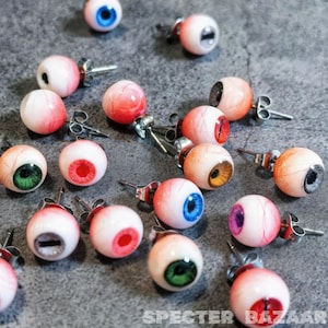 May include: A collection of 16 pairs of eye-shaped earrings with different colored irises. The earrings are made of a white material with a realistic eye design. The earrings are on a gray surface.
