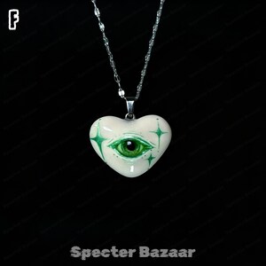 May include: A heart-shaped pendant necklace featuring a glossy off-white surface with a detailed green eye illustration at the center, surrounded by four-pointed green star accents. The pendant hangs from a silver-toned metal chain.