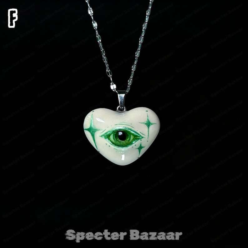May include: A heart-shaped pendant necklace featuring a glossy off-white surface with a detailed green eye illustration at the center, surrounded by four-pointed green star accents. The pendant hangs from a silver-toned metal chain.