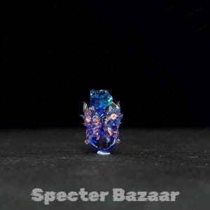 May include: A blue and purple glass sculpture with intricate details. The artwork features a central form with wing-like structures and a textured surface. The base is clear, and the piece is set against a dark background. The text "Specter Bazaar" is at the bottom.