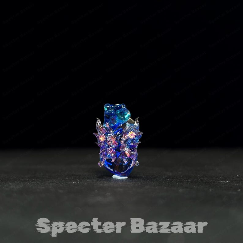 May include: A blue and purple glass sculpture with intricate details. The artwork features a central form with wing-like structures and a textured surface. The base is clear, and the piece is set against a dark background. The text "Specter Bazaar" is at the bottom.