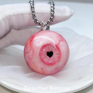 Puede incluir: Un collar con colgante único con un diseño de globo ocular realista. El globo ocular rosa y rojo, con una pupila negra en forma de corazón, cuelga de una cadena plateada. El colgante es una esfera con venas rojas.