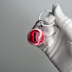 May include: A silver key chain with a red and white eyeball charm. The eyeball has black pupils and a white sclera.