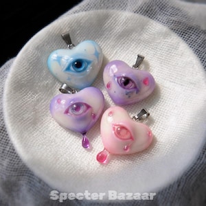 May include: Four heart-shaped pendants with silver-toned bails, each featuring a realistic eye at the center. The pendants are colored in light blue, purple, lavender, and soft pink, with some designs including small teardrop accents.
