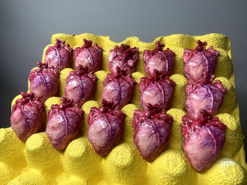 May include: A collection of fourteen small, anatomically detailed heart sculptures arranged in a yellow molded pulp egg carton. Each heart features a glossy, realistic finish with shades of deep red, purple, and pale pink, highlighting veins.