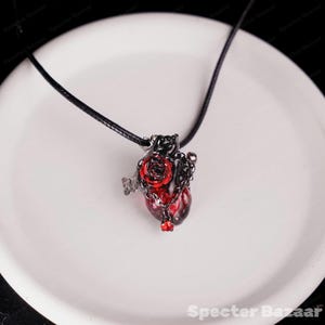 May include: A gothic-style pendant necklace featuring a heart-shaped charm. The charm is a deep red color with a black metal frame, a red rose, and a small chain. The necklace has a black cord.