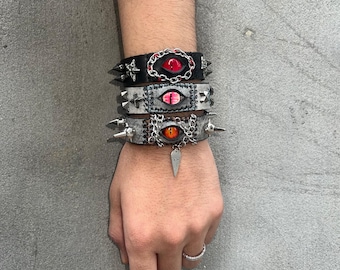May include: Three layered bracelets with silver studs and eye designs. The top bracelet is black leather with a red eye, the middle bracelet is gray leather with a red eye, and the bottom bracelet is gray leather with an orange eye.