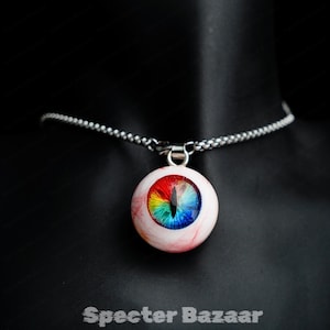 Può includere: Una collana con pendente rotondo con un design a occhio dettagliato. L'occhio ha un'iride arcobaleno e vene rosse, sospeso a una catena d'argento. Il pendente è posto su uno sfondo nero. Il testo "Specter Bazaar" è in basso.