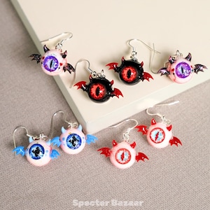 Puede incluir: Una colección de pendientes hechos a mano con un diseño de ojo de diablo. Cada pendiente presenta un ojo redondo con detalles intrincados, pequeños cuernos y alas de murciélago. Los pendientes están disponibles en varios colores, incluyendo rosa, negro y azul, con detalles en rojo y azul.