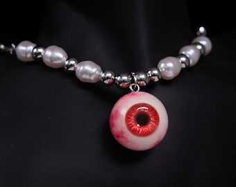 May include: A unique necklace featuring a large, realistic eyeball pendant with a red iris and white sclera. The pendant hangs from a strand of white pearls and silver beads. The necklace is displayed against a black background.