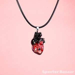 Puede incluir: Un collar con colgante de corazón negro y rojo. El colgante presenta una parte superior en forma de oso negro y una parte inferior en forma de corazón rojo con un diseño de placa de circuito. El collar tiene un cordón negro y un cierre plateado. El fondo es de color rosa liso.
