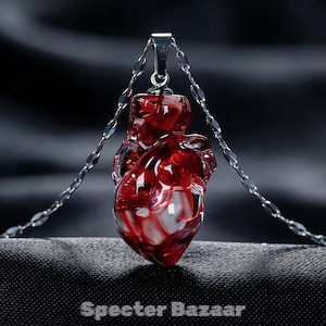 Puede incluir: Un collar con un colgante en forma de corazón rojo translúcido con detalles blancos. El colgante en forma de corazón tiene un diseño detallado. El collar se muestra sobre un fondo oscuro y texturizado.