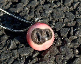 May include: A silver chain necklace with a pink and brown eye pendant. The pendant is round and has two dark brown pupils.