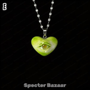 May include: A heart-shaped pendant in shades of lime green and white, featuring a realistic human eye at the center. The pendant hangs from a silver-toned metal chain. The letter E is visible in the top left corner, and the text Specter Bazaar is at the bottom.