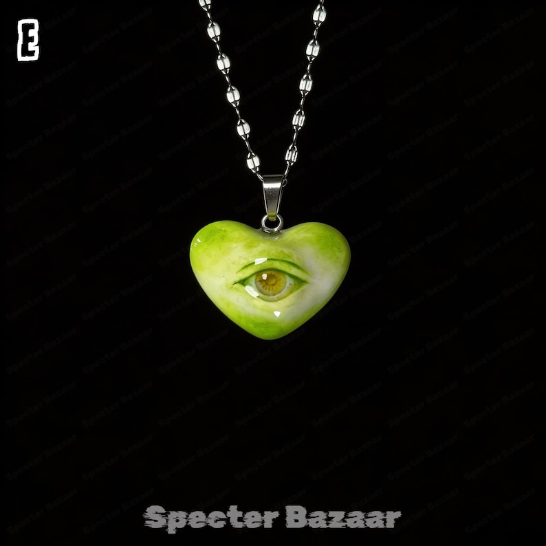 May include: A heart-shaped pendant in shades of lime green and white, featuring a realistic human eye at the center. The pendant hangs from a silver-toned metal chain. The letter E is visible in the top left corner, and the text Specter Bazaar is at the bottom.