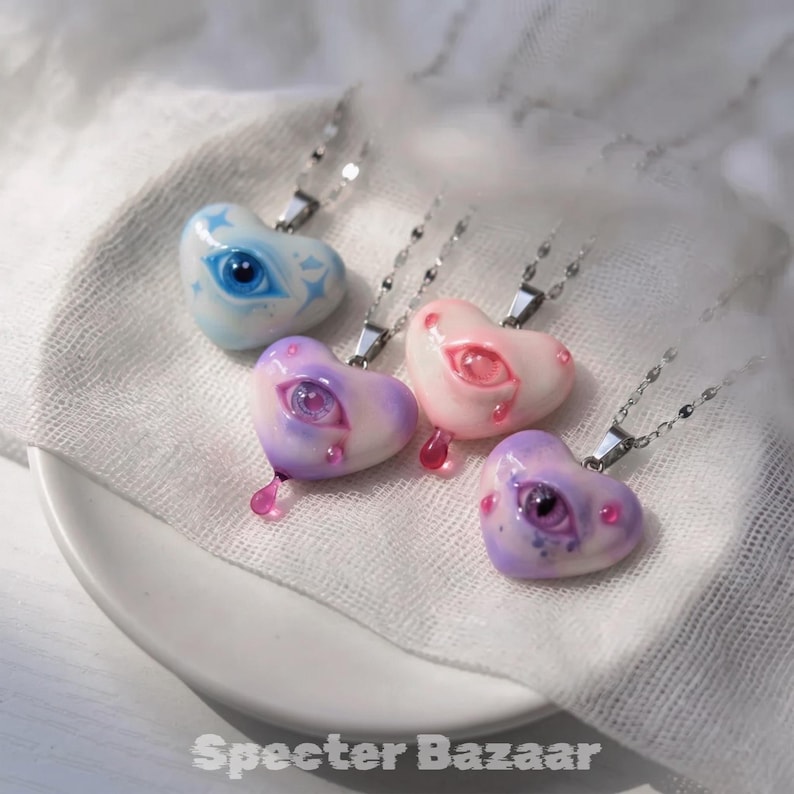 May include: Four heart-shaped pendants on silver-toned chains, each featuring a realistic eye at the center. The pendants are colored in light blue, purple, pink, and lavender, with some showing small teardrop accents and star motifs. Text reads Specter Bazaar.
