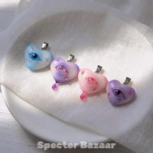 May include: Four heart-shaped pendants with silver-toned bails, each featuring a realistic eye design. The colors are light blue, lavender, pale pink, and deep purple. Two pendants have a small teardrop shape attached to the bottom edge.