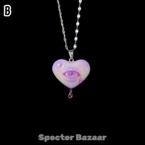 May include: A silver-toned chain necklace featuring a heart-shaped pendant in shades of light purple and white. The pendant displays a central eye graphic with a pink iris and a single pink teardrop, with a small pink droplet hanging from the bottom.