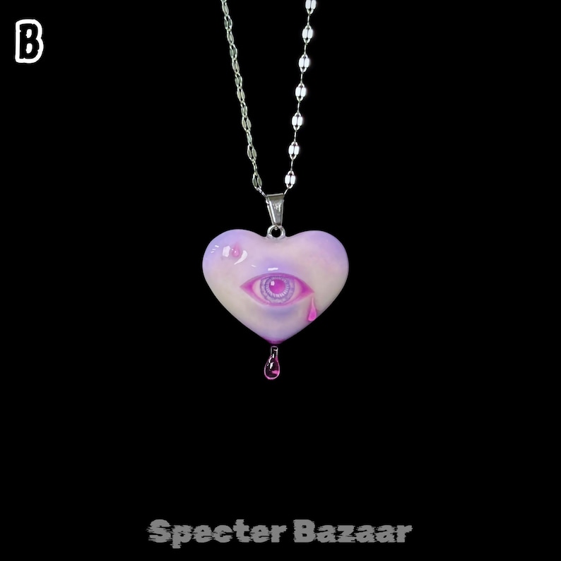 May include: A silver-toned chain necklace featuring a heart-shaped pendant in shades of light purple and white. The pendant displays a central eye graphic with a pink iris and a single pink teardrop, with a small pink droplet hanging from the bottom.