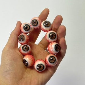 Puede incluir: Un brazalete hecho de múltiples cuentas redondas, cada cuenta está pintada para parecer un globo ocular humano con un esquema de color rojo, blanco y marrón.