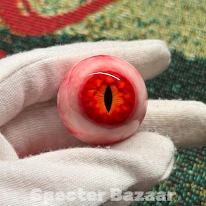 Puede incluir: Un ojo de cristal detallado, con un iris rojo y naranja vibrante y una pupila negra. El ojo está sobre un fondo blanco. El texto "Specter Bazaar" es visible en la parte inferior de la imagen.