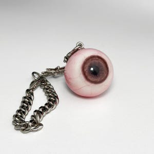 May include: A novelty keychain featuring a realistic eyeball charm. The eyeball is pink with a brown iris and detailed veins. It is attached to a silver-colored chain and metal hardware.