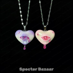 May include: Two heart-shaped pendants on silver-toned chains. One heart is purple and white with a purple eye and teardrop, the other is pink and white with a pink eye and teardrop. Both feature a glossy finish and a central eye design.