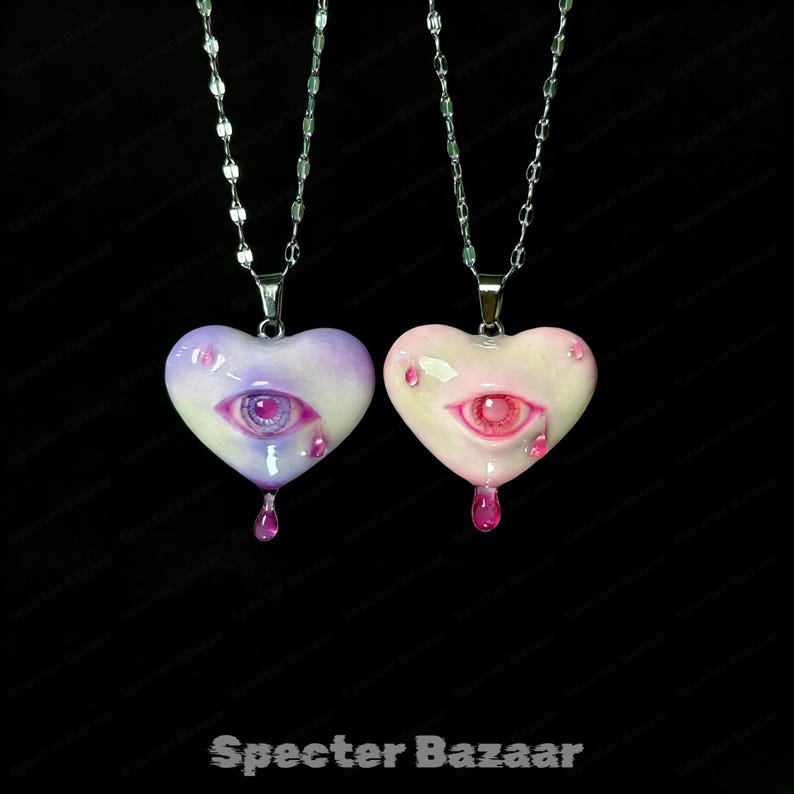 May include: Two heart-shaped pendants on silver-toned chains. One heart is purple and white with a purple eye and teardrop, the other is pink and white with a pink eye and teardrop. Both feature a glossy finish and a central eye design.