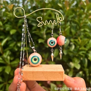 Puede incluir: Un conjunto de joyas con temática de ojos que incluye un collar y pendientes. El collar tiene un colgante con un ojo grande de color rosa con un iris verde y una pupila negra. Los pendientes también son ojos rosas con iris verdes y pupilas negras. Las joyas se muestran en un expositor de madera con la palabra "Smile" escrita en él.