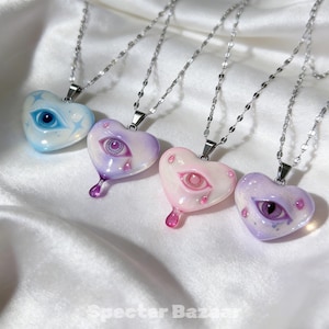 May include: Four heart-shaped pendants on silver-toned chains, each featuring a central eye illustration. The pendants are light blue, lavender, pale pink, and purple, with some designs including small teardrop accents and star motifs.