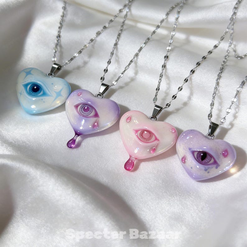 May include: Four heart-shaped pendants on silver-toned chains, each featuring a central eye illustration. The pendants are light blue, lavender, pale pink, and purple, with some designs including small teardrop accents and star motifs.
