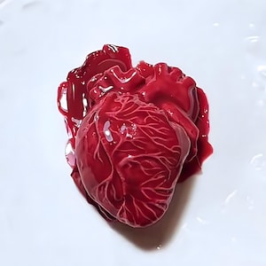 May include: A realistic anatomical heart model made of red resin. The heart is detailed with veins and chambers, and is sitting on a white surface.
