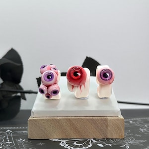 Puede incluir: Tres anillos con diseños de ojos realistas. El anillo izquierdo tiene múltiples iris morados pequeños sobre una banda blanca. El anillo central presenta un solo ojo grande de color rojo con venas visibles. El derecho muestra un ojo con iris morado.