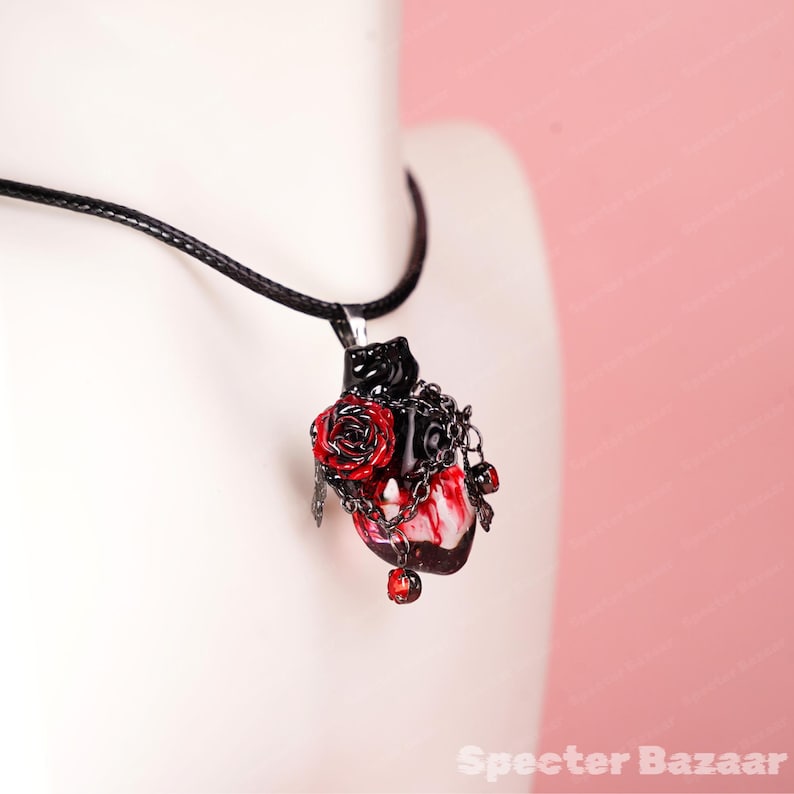 May include: A gothic-style pendant necklace featuring a heart-shaped charm with a black and red color scheme. The charm includes a red rose, chains, and red bead accents, suspended from a black cord.