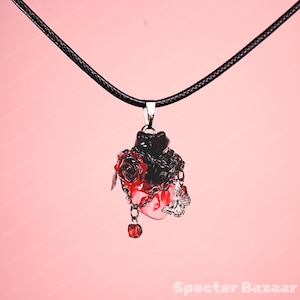 May include: A black pendant necklace with a heart-shaped design. The pendant features a black cat, red roses, and silver chains. A small silver butterfly charm and a red gem dangle from the bottom. The necklace hangs on a black cord.