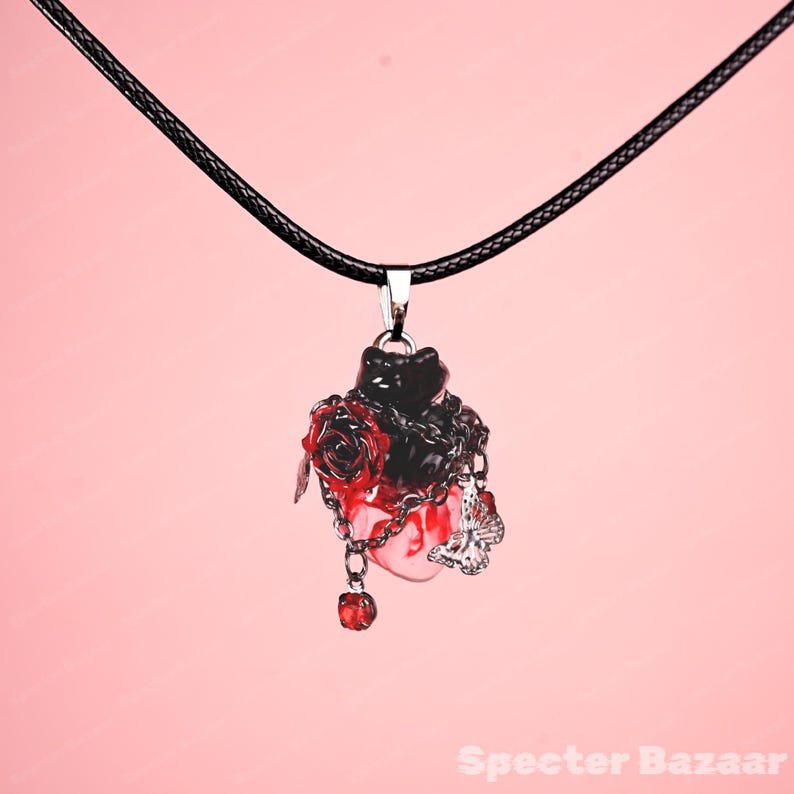 May include: A black pendant necklace with a heart-shaped design. The pendant features a black cat, red roses, and silver chains. A small silver butterfly charm and a red gem dangle from the bottom. The necklace hangs on a black cord.