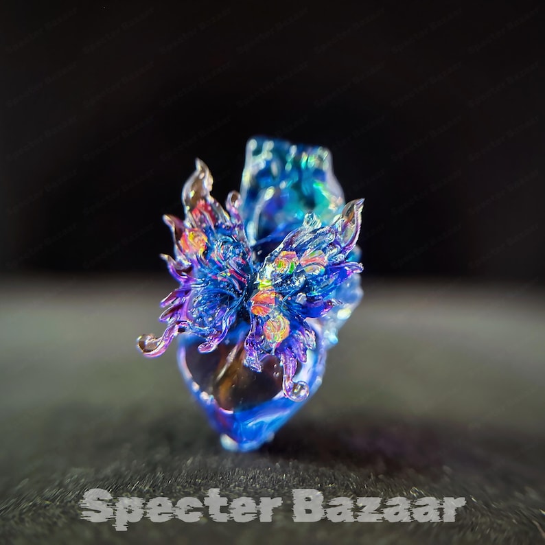 May include: A glass pendant featuring a heart-shaped base in shades of deep blue and purple, topped with intricate, flame-like glass filigree in iridescent blue, violet, and orange hues. The piece displays a glossy, multi-dimensional finish.